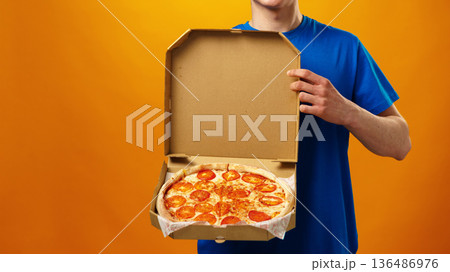 Man holds pizza box with pepperoni pizza against orange background at delivery time Man holds pizza box with pepperoni pizza against orange background at delivery time 136486976