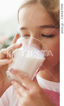 Girl drinks milk from a glass while sitting indoors during daylight Girl drinks milk from a glass while sitting indoors during daylight 136487006
