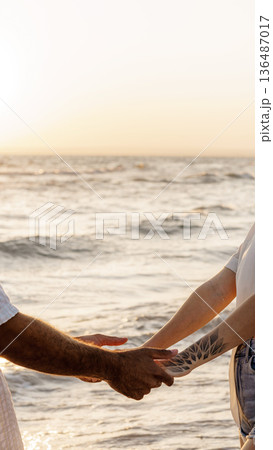 Couple holds hands at the beach during sunset with ocean waves in the background Couple holds hands at the beach during sunset with ocean waves in the background 136487017