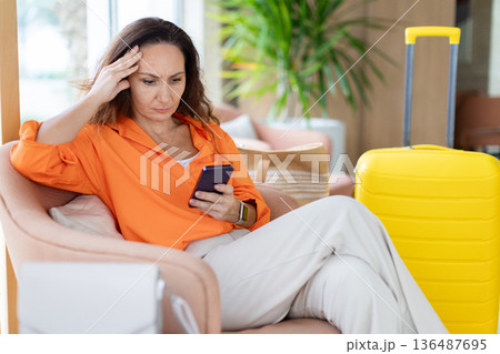 Adult woman sitting in hotel lobby holding smartphone and touching her head 136487695