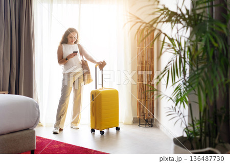 Woman standing by window checking smartphone with yellow suitcase in hotel room Woman standing by window checking smartphone with yellow suitcase in hotel room 136487730