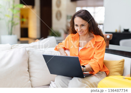 Adult woman using laptop on hotel sofa, remote work during travel stay 136487734