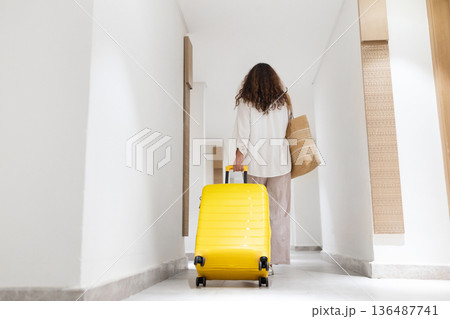 Adult woman walking away through long hotel hallway with suitcase, minimalist interior Adult woman walking away through long hotel hallway with suitcase, minimalist interior 136487741