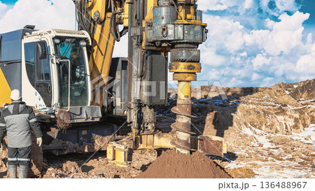A drilling rig is actively working on a pile foundation at a construction site. A worker is present near the rig observing the operation 136488967