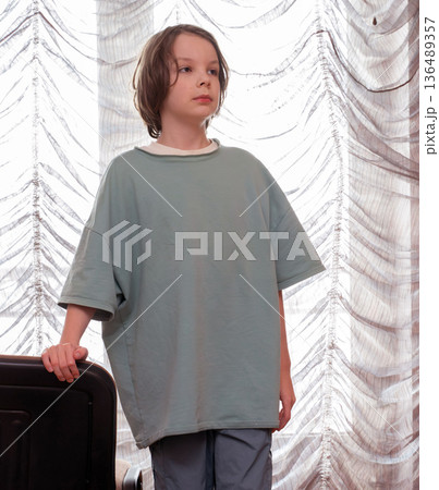 Vertical portrait of a pensive boy in an oversized t-shirt by a window. Child at home 136489357