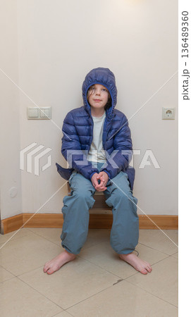 Young boy in a blue hooded down jacket sitting barefoot in a corner. Winter coat indoors 136489360