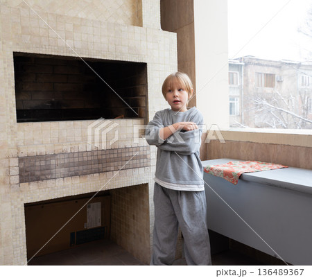 Young boy standing on a balcony next to a fireplace. Child feeling cold in winter Young boy standing on a balcony next to a fireplace. Child feeling cold in winter 136489367