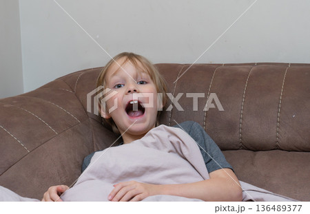 Tired young boy yawning with mouth wide open on a sofa. Sleepy child waking up at home 136489377