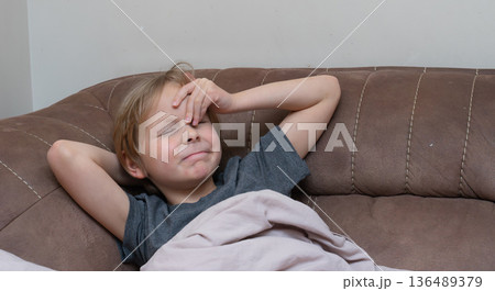 Young boy with a headache touching his forehead on a sofa. Sick child feeling unwell at home Young boy with a headache touching his forehead on a sofa. Sick child feeling unwell at home 136489379