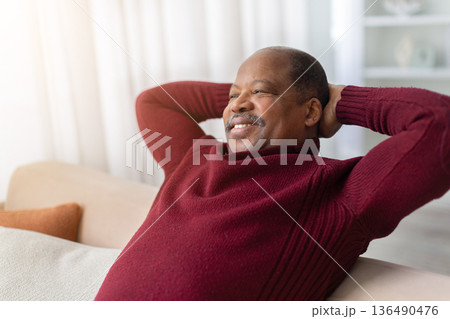 Relaxed black man sitting on sofa at home with hands behind head 136490476