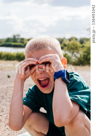 Young boy squints his eyes while making hand binoculars and laughing. He wears a green shirt and a blue smartwatch Young boy squints his eyes while making hand binoculars and laughing. He wears a green shirt and a blue smartwatch 136491042