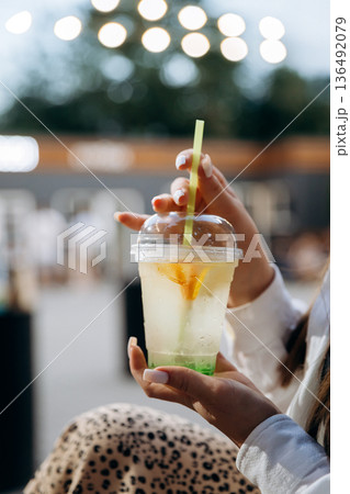 Woman holding iced lemon drink in plastic cup with straw, outdoor cafe bokeh 136492079
