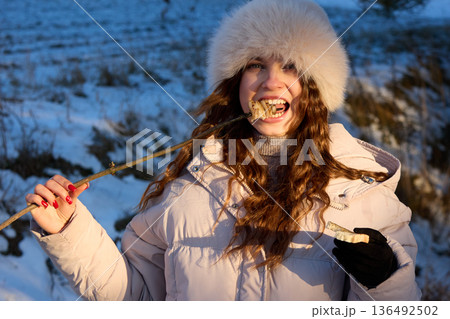 Happy woman in a fur hat eats roasted marshmallows on a stick outdoors in winter. Happy woman in a fur hat eats roasted marshmallows on a stick outdoors in winter. 136492502
