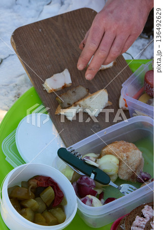 Picnic outdoors: A hand places bacon on bread, amidst food containers and a snowy landscape. 136492629