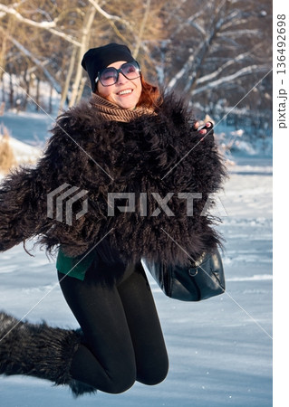 A joyful woman in a stylish fur coat jumps for joy in a snowy winter landscape in a good mood. 136492698