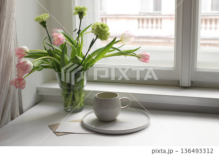Easter spring still life. Cup of tea coffee and floral bouquet on window sill. Pink tulips, green carnation flowers in vase. Blank greeting card mockup. Home decor, breakfast. Scandinavian interior Easter spring still life. Cup of tea coffee and floral bouquet on window sill. Pink tulips, green carnation flowers in vase. Blank greeting card mockup. Home decor, breakfast. Scandinavian interior 136493312