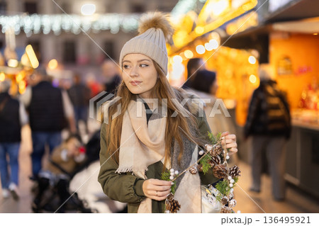 Girl with cone and fir brunch garland stands in middle fair decorated for New Year holidays Girl with cone and fir brunch garland stands in middle fair decorated for New Year holidays 136495921
