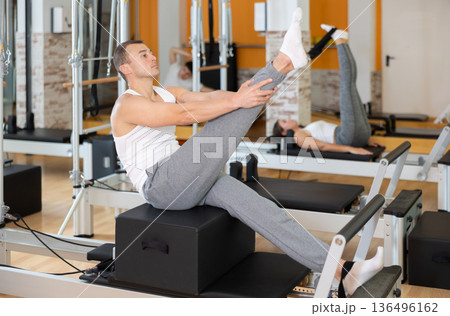 Man works out in gym and performs set of exercises to stretch leg muscles with help of reformer 136496162