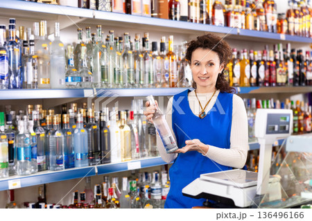 Woman salesperson in alcohol department of Russian store stand with vodka near showcase. Woman salesperson in alcohol department of Russian store stand with vodka near showcase. 136496166
