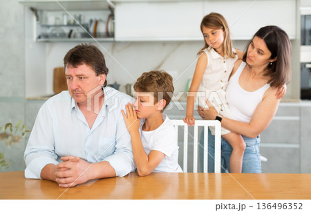 Frustrated man sits at kitchen table and tells wife and children about unforeseen difficulties 136496352