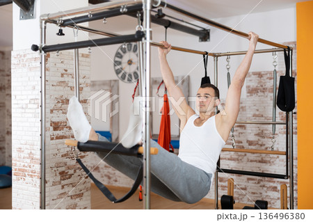 Man visitor performs exercises in hanging position on combined Pilates trapeze table. 136496380