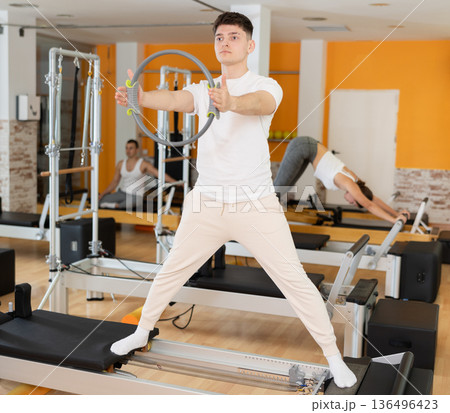 Man stands on reformer and holds ring, hoop in his hands, performs exercises to balance 136496423