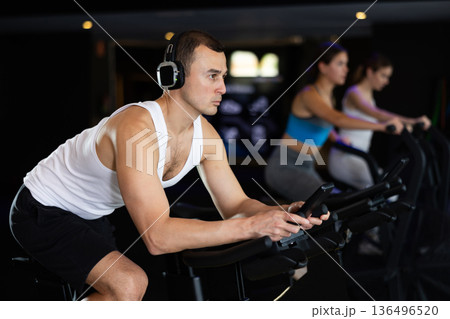 Man in earphone performs exercises with stationary bicycle in gym Man in earphone performs exercises with stationary bicycle in gym 136496520