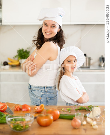 Mother and girl in white cooking hats are standing in kitchen near table 136496538