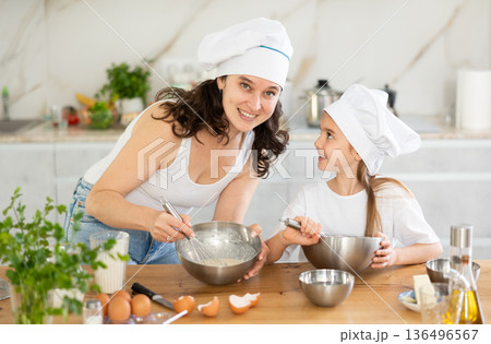 Mom and girl in white chief hats are talking in kitchen and whisking ingredients for pastry in bowl Mom and girl in white chief hats are talking in kitchen and whisking ingredients for pastry in bowl 136496567