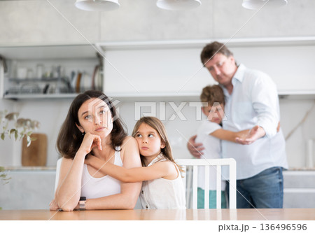 Offended mother sitting at the kitchen table being hugged by little daughter 136496596