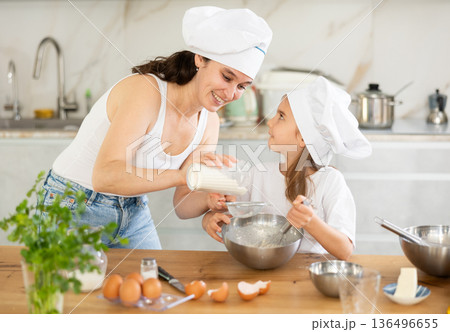Mom sifts and adds flour to bowl, girl mixes ingredients with whisk. Mom sifts and adds flour to bowl, girl mixes ingredients with whisk. 136496655