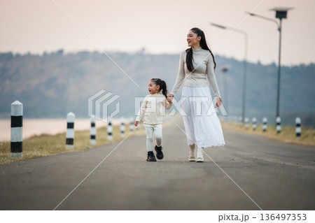 Mother and child girl holding hands walking on road by the lake at sunset. 136497353