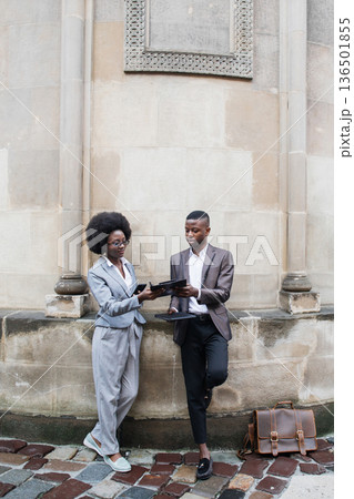 Two smartly dressed African American business professionals, a woman and a man, are engaged in a discussion while holding tablets outside a stone building Two smartly dressed African American business professionals, a woman and a man, are engaged in a discussion while holding tablets outside a stone building 136501855