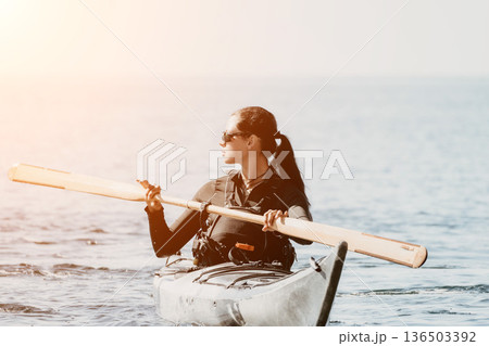 Kayaking Woman Paddle Ocean - A woman in a kayak on the ocean holding a paddle with the sun setting in the background. 136503392