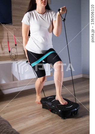 Brunette girl exercises with expanders at home on a vibration platform to reduce weight and cellulite. Exercise at home, lifestyle Brunette girl exercises with expanders at home on a vibration platform to reduce weight and cellulite. Exercise at home, lifestyle 136506110