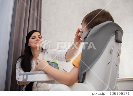 Mother feeds child sitting in high chair, child watches tablet, everyday activity of feeding time, focus on family care, comfortable indoor scene 136506171