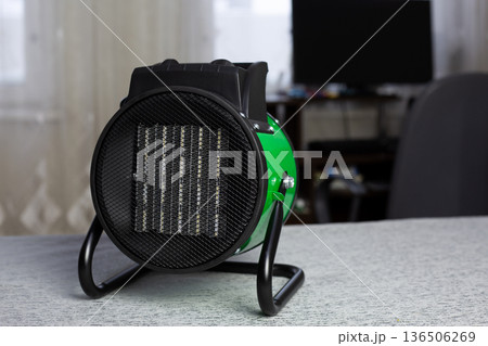 Heat gun against desk with computer in home room. Concept for raising temperature in room during cold season. Room heating device against the background of living interior Heat gun against desk with computer in home room. Concept for raising temperature in room during cold season. Room heating device against the background of living interior 136506269