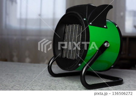 Green heater on stand against window background. Compact ceramic space heater with fine mesh front and handle on top. Room heater. Raising temperature in cold room with heater 136506272