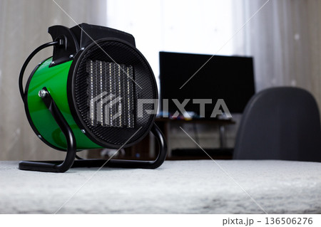 Heat gun with radiator and fan for warming room. Green electric heater for increasing air temperature in room. Compact cylindrical heater in interior. How to warm house in winter 136506276