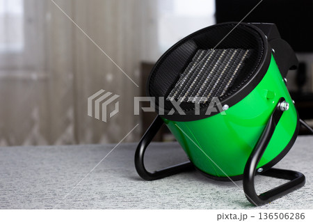 Raising the room temperature. Electric round heater with radiator and fan for circulating warm air throughout the room. Heating the room for living. Green heat gun on stand is pointed upwards Raising the room temperature. Electric round heater with radiator and fan for circulating warm air throughout the room. Heating the room for living. Green heat gun on stand is pointed upwards 136506286