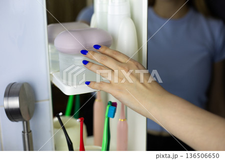 Girl hand reaches for cleaning products in bathroom near mirror. Skincare products on shelf in lived-in bathroom. Natural interior with shelves of hygiene products, Girl routine begins Girl hand reaches for cleaning products in bathroom near mirror. Skincare products on shelf in lived-in bathroom. Natural interior with shelves of hygiene products, Girl routine begins 136506650