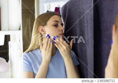 Young woman with blond hair performs home beauty routine in front of mirror. Portrait of woman applying moisturizing cream to her face. Woman is taking care of her skin youthfulness and freshness Young woman with blond hair performs home beauty routine in front of mirror. Portrait of woman applying moisturizing cream to her face. Woman is taking care of her skin youthfulness and freshness 136506711