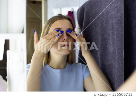 Young woman massages her eyelids with special moisturizer. Skin care and facial wrinkle prevention with moisturizing massage. Young woman skin care and beauty routine in bathroom Young woman massages her eyelids with special moisturizer. Skin care and facial wrinkle prevention with moisturizing massage. Young woman skin care and beauty routine in bathroom 136506712