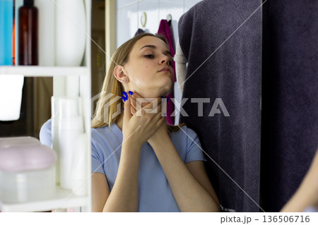 Moisturizing woman neck in front of bathroom mirror. Woman rubs care product into her skin for healthy, fresh skin. Taking care of your body through face cream. Neck skin health concept 136506716