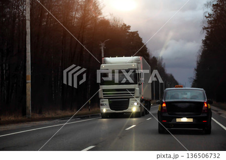 Nighttime road scene with semi-trailer truck driving towards camera on highway while black taxi car travels ahead, showing logistics and freight delivery. Copy space for text Nighttime road scene with semi-trailer truck driving towards camera on highway while black taxi car travels ahead, showing logistics and freight delivery. Copy space for text 136506732