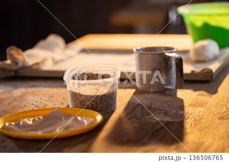 Kitchen table scene shows food preparation ingredients with granular seeds in plastic container, measuring cup, and yellow plate holding light-colored dough or paste, featuring bright soft light Kitchen table scene shows food preparation ingredients with granular seeds in plastic container, measuring cup, and yellow plate holding light-colored dough or paste, featuring bright soft light 136506765