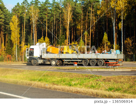 White semi truck with flatbed trailer transporting yellow construction equipment, including small loader, parked on roadside next to autumn forest with green pine and yellow birch trees on sunny day White semi truck with flatbed trailer transporting yellow construction equipment, including small loader, parked on roadside next to autumn forest with green pine and yellow birch trees on sunny day 136506914