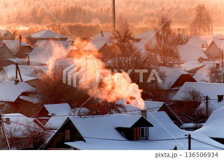 Golden sunrise light illuminating smoke plume rising from chimney of house with snow-covered roof in tranquil frosty winter village morning scene Golden sunrise light illuminating smoke plume rising from chimney of house with snow-covered roof in tranquil frosty winter village morning scene 136506934