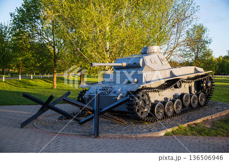 Historical military tank monument displayed in a public park setting during golden hour, featuring a grey battle tank and anti-tank obstacles surrounded by green trees and grass Historical military tank monument displayed in a public park setting during golden hour, featuring a grey battle tank and anti-tank obstacles surrounded by green trees and grass 136506946