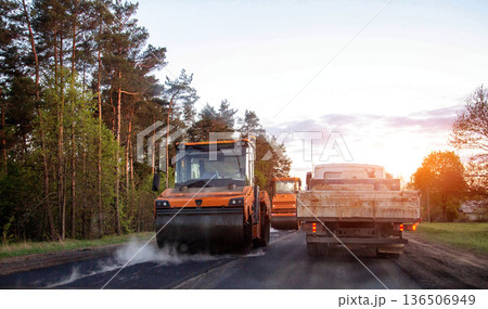 Road construction scene with heavy machinery compacting fresh asphalt surface on rural road at sunset, forest edge background and warm light Road construction scene with heavy machinery compacting fresh asphalt surface on rural road at sunset, forest edge background and warm light 136506949